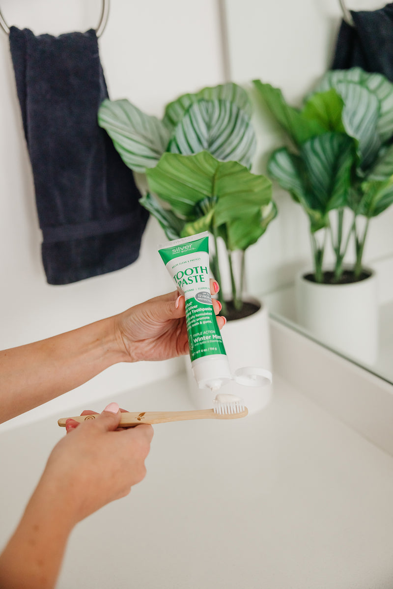 Person holding a tube of toothpaste and a toothbrush in a bathroom setting with plants in the background.